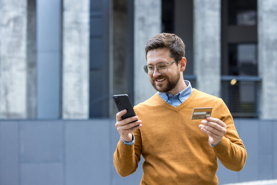 Close-up photo of a young man standing outside, holding a credit card and using a mobile phone