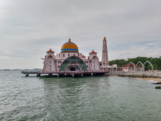 Fototapeta premium Scenic view of iconic white Malacca Straits Mosque stands partially in water against vibrant sky in Malaysia, Southeast Asia. Golden dome and tall minaret are prominent. Religious muslim landmark