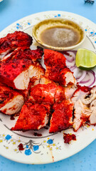 Plate of tandoori chicken, cut into pieces, is presented with a small bowl of sauce and a lime wedge. The chicken has a vibrant reddish-orange color from the tandoori marinade. Red onion slices