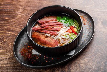 A close-up of a flavorful beef noodle soup served in a black bowl. The dish features tender slices of beef, fresh herbs, and red chili, all presented on a wooden table.