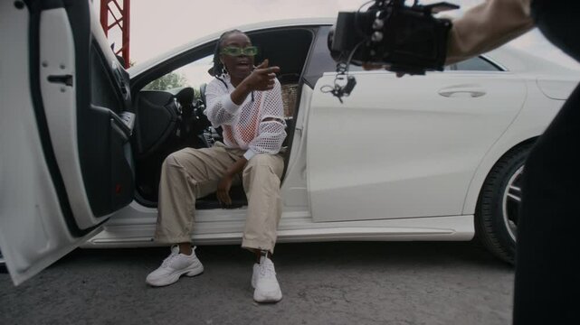 Full shot of music video recording process - male cameraman filming young Black female hip-hop artist in classy outfit and glasses sitting in white car and rapping to camera
