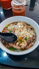 White bowl of beef ho fun noodle soup is the focus. Wide, flat rice noodles (ho fun) swim in a dark broth with slices of beef and chopped green onions. Southeast Asia cuisine. Street food market