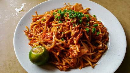Plate of mie goreng fried noodles. Coated in a reddish-orange sauce and garnished with chopped green onions. Lime wedge sits on the side of white plate. Southeast Asian cuisine. Street food market