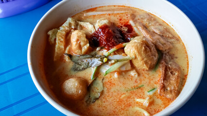 Bowl of curry mee Laksa, spicy noodle soup. Creamy, orange-tinged broth contains yellow noodles, tofu puffs, fish balls, and other ingredients like vegetables and dollop of chili paste. Rich flavorful
