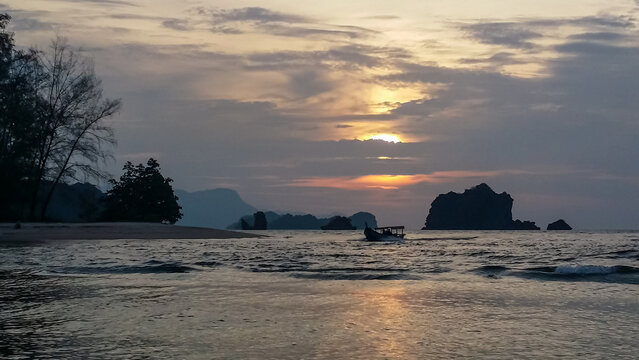 Tranquil coastal scene at dusk or dawn in Kilim Geoforest Park, Langkawi Island, Malaysia. sky is filled with dramatic, layered clouds. Gentle waves lap sandy shore. Dark rocky outcrops rise from sea