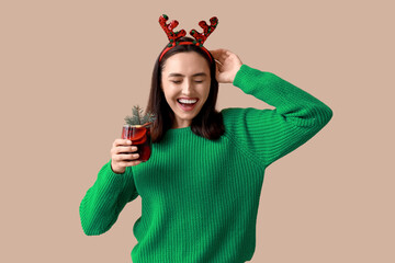 Happy young woman in reindeer horns with glass of hot mulled wine on brown background
