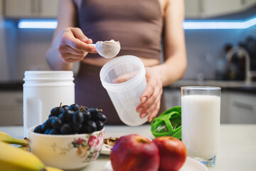 Athletic woman in sportswear preparing protein drink in a kitchen, putting a portion of whey protein powder from a measuring spoon into a shaker in her hand