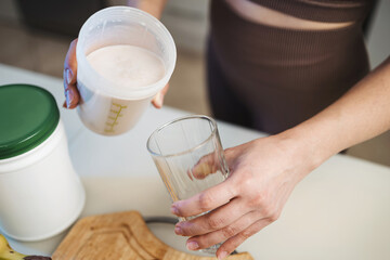 Athletic woman in sportswear pouring a protein drink, milkshake, or smoothie from a shaker into a glass in her hand, in the kitchen	