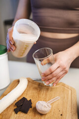 Athletic woman in sportswear pouring a protein drink, milkshake, or smoothie from a shaker into a glass in her hand, in the kitchen