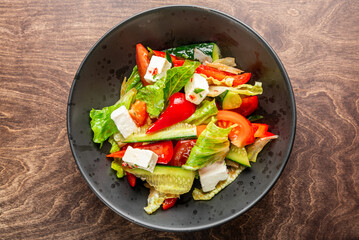 A fresh, colorful salad featuring crisp lettuce, ripe tomatoes, crunchy cucumbers, vibrant bell peppers, and creamy feta cheese, served in a sleek black bowl on a wooden table.
