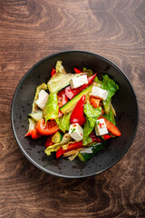 A fresh, colorful salad featuring crisp lettuce, ripe tomatoes, crunchy cucumbers, vibrant bell peppers, and creamy feta cheese, served in a sleek black bowl on a wooden table.