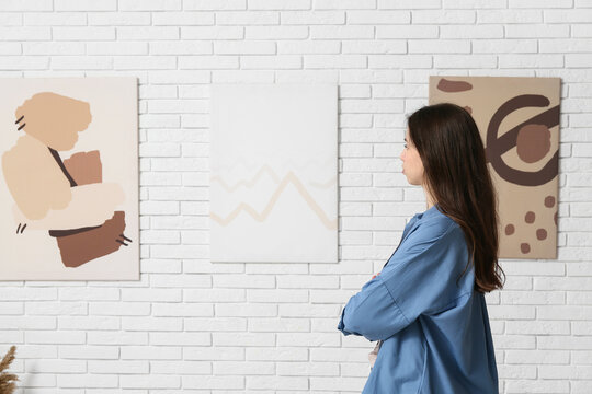 Young woman looking at paintings on white brick wall in art gallery