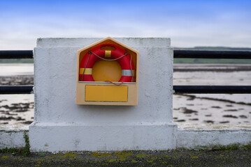 A vibrant red and yellow lifebuoy on a concrete wall beside coastal railings, overlooking serene...