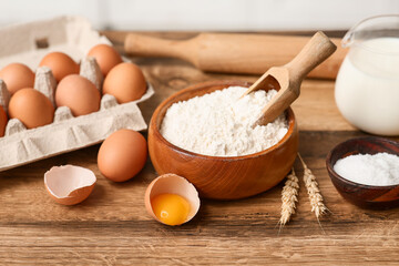 Bowl of flour with scoop, eggs, jug of milk and salt for preparing dough on kitchen counter
