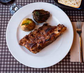 Delicious grilled beef ribs served on plate with side dish of boiled potatoes and baked artichokes. Popular Spanish combination meal