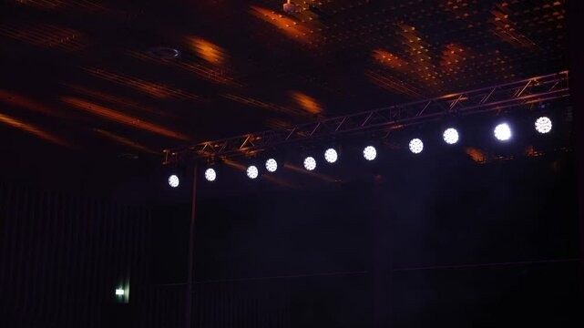 Row of bright white spotlights on a truss in an indoor venue, with a patterned ceiling and warm lighting reflections. An exit sign is visible on the left.