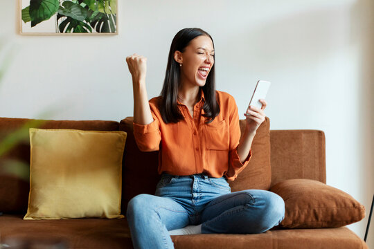 Euphoric lady getting good news notification via smartphone, making yes gesture and shouting, celebrating success while sitting on couch at home - Powered by Adobe