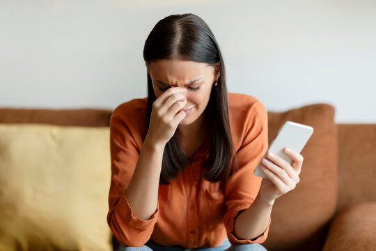 Worried lady holding smartphone and touching nose bridge, frustrated woman reading unpleasant sms, emotionally reacting to bad message and crying