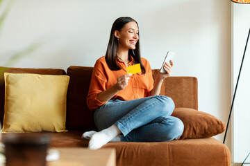 Online transaction concept. Positive woman holding smartphone and credit card for payment, online shopping or transaction, sitting on couch at home