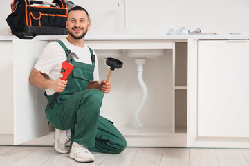Male plumber with pipe wrench and plunger near sink in kitchen