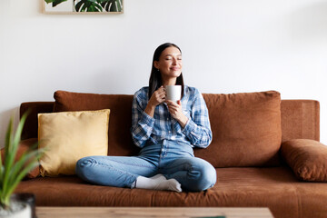 Smiling relaxed woman enjoying the warmth of cup of coffee, sitting cross-legged on comfy sofa in bright, contemporary living room
