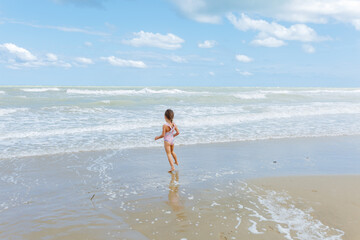 rea view of little girl in the swimsuit jumping on the beach playing with waves