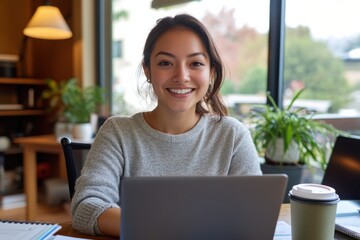 A happy young woman smiles while using her laptop, with a cup of coffee beside her in a bright space, radiating positivity and the joy of productivity in a cozy setting.