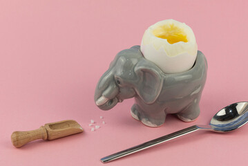 Das perfekt weich gekochte Ei in einem lustigen Eierbecher in Tierform (Elefant) mit Salz und Eierlöffel auf einem rosafarbenen flachen Hintergrund für Freude und Spaß beim frühstücken, Platz für Text