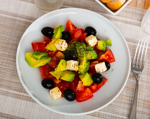 Fresh avocado salad with tomatoes, black olives and feta cheese served in bowl with serving pieces.