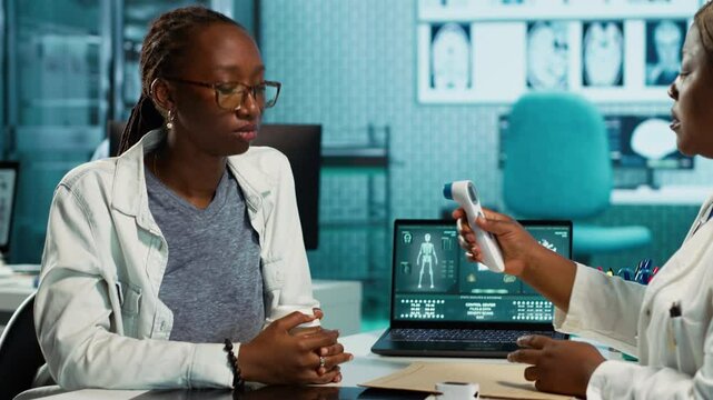 Black medic measuring the temperature during routine checkup at the clinic, using a digital thermometer to check for flu symptoms. Health specialist ensures diagnosis for a patient. Camera A.