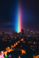 A vibrant, vertical rainbow beam illuminates a cityscape at night, casting colorful light over tall buildings and streets below.