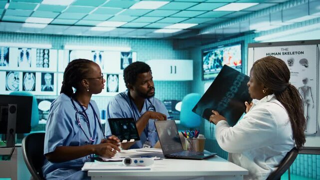 African american general practitioner leading a healthcare team in a cabinet, analyzing MRI imaging results and providing accurate diagnosis and effective treatment. Medical facility. Camera B.