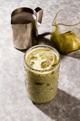 Iced Matcha Latte served into cold glass with ice, an empty glass jar, an a milk silver jar over a grey background