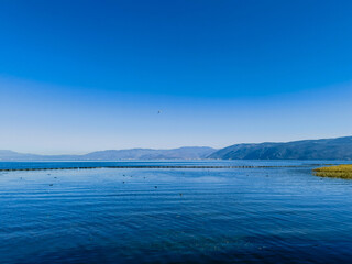 Serene lake scene under a vibrant blue sky.  Calm water reflects the distant mountains, creating a tranquil atmosphere.  A hint of shoreline vegetation is visible.