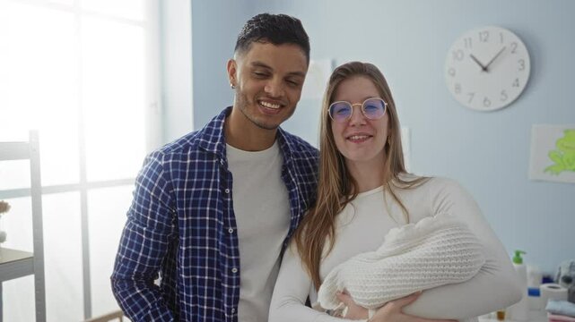 Couple happily embracing their newborn in a cozy bedroom, capturing a joyful family moment indoors with a modern clock on the wall.