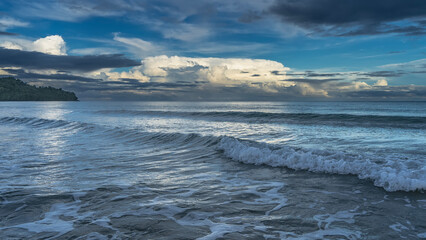Obraz premium Morning. The clouds in the blue sky are highlighted in gold. The ocean waves roll towards the shore, foam, spread out. Glare on the sea surface. A hill in the distance. Malaysia. Borneo. Kota Kinabalu