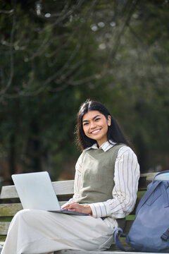 Vertical portrait of happy Asian teen girl college student on bench in nature park using computer device studying online hybrid learning looking at camera sitting with laptop.