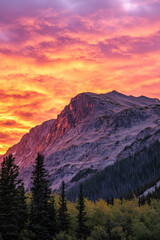 Obraz premium A breathtaking sunset illuminates the rocky mountain peak with vibrant orange and pink hues, surrounded by lush evergreen and deciduous trees.