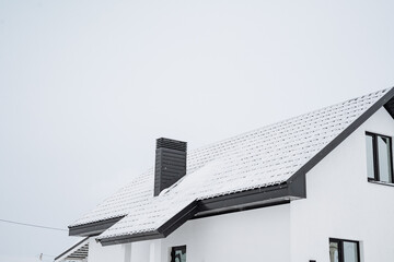 The roof of a beautiful white house is currently covered in a thick layer of snow that has fallen recently, creating a picturesque winter scene that showcases the charming architecture