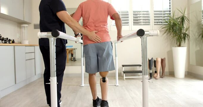 Therapist assisting man with prosthetic leg walking on parallel bars indoors
