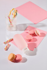 High-angle view of a cosmetics ad photo. The dessert table theme shows pink macarons, candy canes in a clear glass, pink paper cards, and a pale pink cosmetic bottle mockup resting on a white surface.