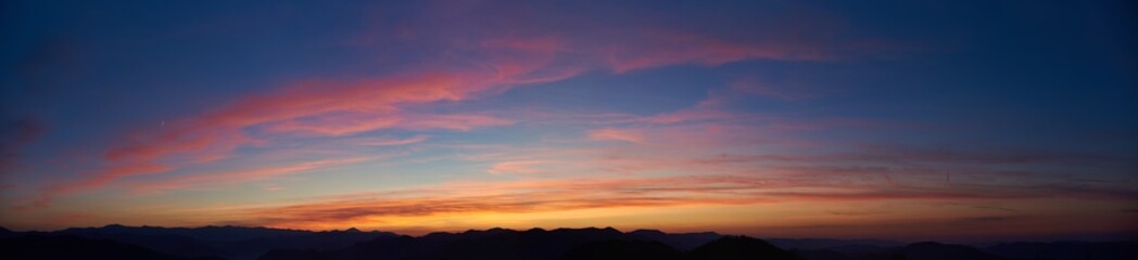 Obraz premium Mesmerizing sunset paints sky with vibrant hues of pink, orange, and blue. Soft, wispy clouds stretch across horizon, while silhouettes of distant mountains create dramatic and tranquil scene.