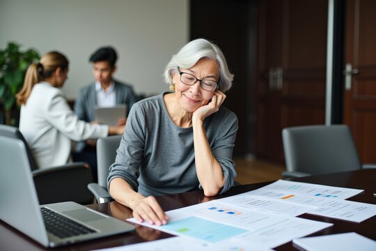 Senior Businesswoman Analyzing Financial Reports in Modern Office Setting with Colleagues Collaborating in the Background - Powered by Adobe