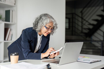 Elderly businesswoman calculating figures while engaged with a laptop in her modern office