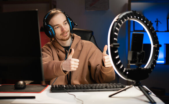 blogger young gamer broadcasts live streaming to review video game on social media, showing thumbs-up, reviews for video blog. Influencer uses smartphone and ring lamp