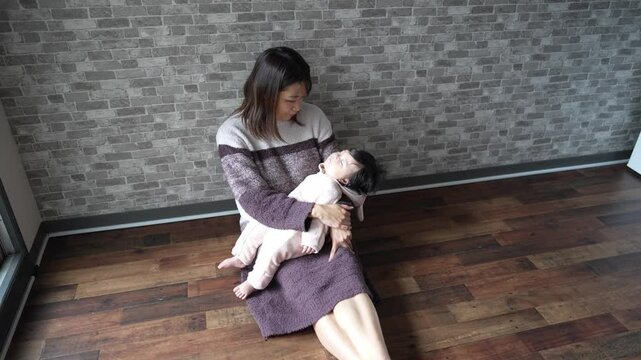 In a warm winter Japanese apartment room, a 6-month-old Japanese girl wearing fluffy clothes and her mother are sitting on the floor holding their sleeping baby.