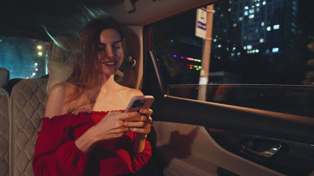 A young red-haired woman dressed elegantly in a stunning red dress is using her smartphone while comfortably seated in a luxurious car at night