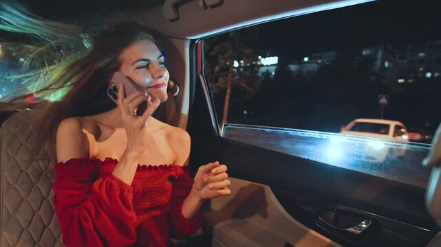 A young red-haired woman dressed elegantly in a stunning red dress is using her smartphone while comfortably seated in a luxurious car at night