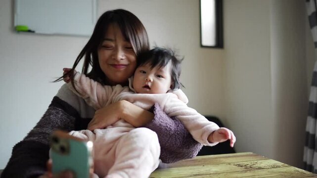 A 6-month-old Japanese girl and her mother wearing fluffy clothes in a warm winter Japanese apartment room. Baby sitting at desk and mother looking at smartphone.