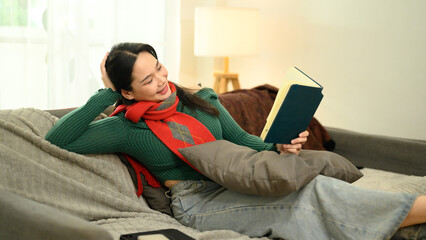 Cheerful young woman in green sweater and red scarf reading a book on couch